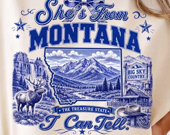 May include: A cream-colored t-shirt featuring a blue graphic print. The text reads "She's From MONTANA" and "I Can Tell" with "THE TREASURE STATE" on a banner. The design includes a mountain landscape, an elk, a cabin, and a cowboy hat.
