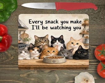 May include: A rectangular cutting board featuring an illustration of eight cats gathered around a food bowl on a wooden counter. The text reads, Every snack you make I'll be watching you. The board is positioned near a red-handled knife and vegetables.