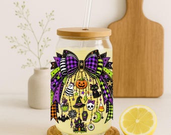 May include: A clear glass tumbler with a bamboo lid and a glass straw, featuring a colorful Halloween-themed graphic of a large purple plaid bow adorned with hanging charms including a pumpkin, ghost, black cat, witch hat, skull, and haunted house.