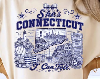 May include: A cream-colored t-shirt featuring a blue graphic print with the text "She's CONNECTICUT I Can Tell." The design includes a map of Connecticut, a lighthouse, a sailboat, a beach bag, a wooden sign listing cities, and a bow at the top.