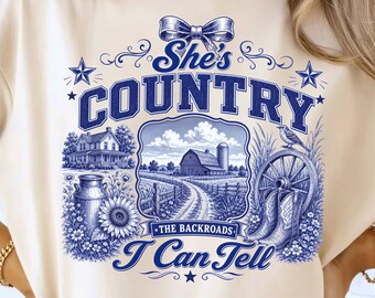 May include: A cream-colored t-shirt featuring a blue graphic print with the text "She's COUNTRY I Can Tell" and "THE BACKROADS". The design includes a farmhouse, a barn, a milk can, sunflowers, a wagon wheel, cowboy boots, and a bird.