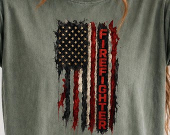 May include: A sage green crew neck t-shirt featuring a distressed, vertical American flag graphic. The design incorporates black, white, and red colors with the word FIREFIGHTER printed in bold, vertical red capital letters along the right side.