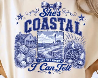 May include: A cream-colored t-shirt featuring a blue graphic print with the text She's COASTAL THE SEASHORE I Can Tell. The design includes a lighthouse, a sailboat, a house, seashells, a seagull, a wooden wheel, and a decorative bow.