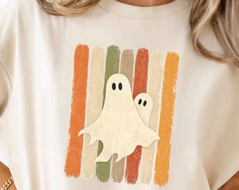 May include: A cream-colored t-shirt featuring a graphic of two off-white ghosts centered over a series of vertical, distressed stripes in shades of orange, yellow, and muted green. The ghosts have simple black oval eyes and a textured appearance.