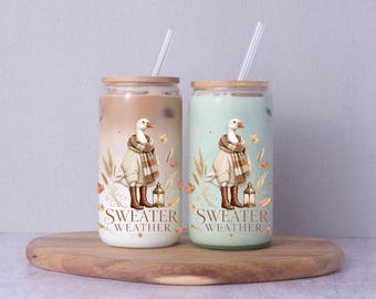 May include: Two glass tumblers with bamboo lids and clear straws, each filled with a different colored beverage. The glasses feature a graphic of a goose wearing a sweater and boots, surrounded by wheat and a lantern, with the text SWEATER WEATHER.