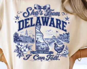 May include: A cream-colored t-shirt featuring a blue graphic print with the text She's From DELAWARE, THE FIRST STATE, and I Can Tell. The design includes a map of Delaware, a lighthouse, a chicken, flowers, and a historic building.