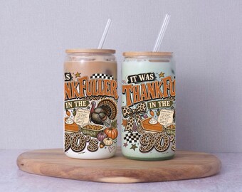 May include: Two glass tumblers with bamboo lids and clear straws, each featuring a graphic print with the text IT WAS THANKFULLER IN THE 90'S, a turkey, pumpkin pie, a leopard print bow, and a checkered pattern in orange, brown, and green tones.