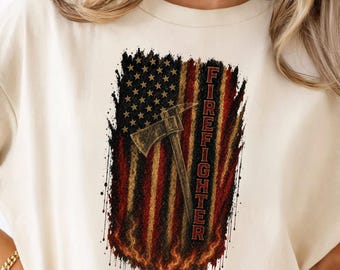 May include: A cream-colored t-shirt featuring a graphic print of a distressed American flag with a fire axe overlaid in the center. The word FIREFIGHTER is printed vertically in red block letters along the right side of the flag design.