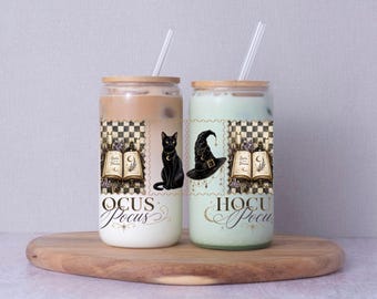 May include: Two clear glass tumblers with bamboo lids and clear straws, filled with light brown and light green beverages. The glasses feature a graphic print of a black cat, a witch hat, and an open book with the text Hocus Pocus.
