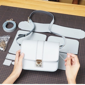 Pode incluir: Um padrão de bolsa de couro azul claro com um fecho de metal. O padrão inclui as peças da bolsa, da alça e da aba. O padrão está disposto em uma grade cinza.