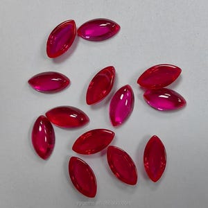 May include: A selection of marquise-cut, faceted gemstones in shades of red and pink. The gems are scattered on a light-coloured surface. The image includes the website address "yygems.en.alibaba.com".
