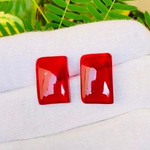 Puede incluir: Dos piedras preciosas rectangulares rojas sobre una superficie blanca. Las piedras preciosas son lisas y brillantes.