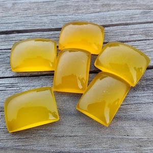 May include: Six yellow, rectangular, translucent cabochons, likely for jewelry making, are arranged on a wooden surface.