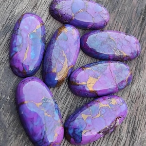 May include: Seven oval-shaped gemstones with a polished finish. The stones feature a mix of purple, blue, and gold colors. The gemstones are arranged on a wooden surface, showcasing their unique patterns and textures. Ideal for jewelry making.