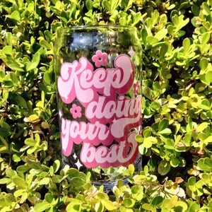 May include: A pink glass with the text "Keep doing your best" in a retro font. The glass is surrounded by green foliage.