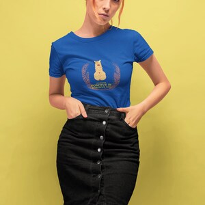 Op de afbeelding: Een vrouw draagt een koninklijk blauw t-shirt met een lama-afbeelding en de tekst "Wees altijd positief op een positieve manier". Ze draagt een zwarte denim rok en heeft rood haar.
