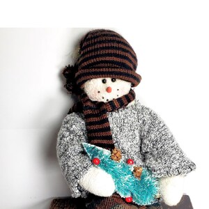 Op de afbeelding: Een pluche sneeuwpoppop met een bruin-zwarte gestreepte muts en sjaal, die een kleine groene kerstboom vasthoudt. De sneeuwpop zit op een houten kist.