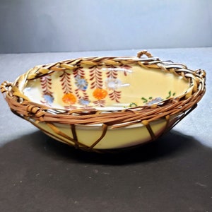 Puede incluir: Un cuenco decorativo de cerámica con un diseño de cesta de mimbre tejida. El cuenco tiene un interior amarillo pálido con patrones florales en naranja, azul y marrón. El exterior es una cesta de mimbre marrón tejida.