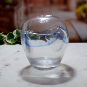 Op de afbeelding: Heldere glazen vaas met een afgeronde vorm en een kleine opening. Het heeft een blauw wervelpatroon in het midden. Het glas heeft een lichte iriserende glans.