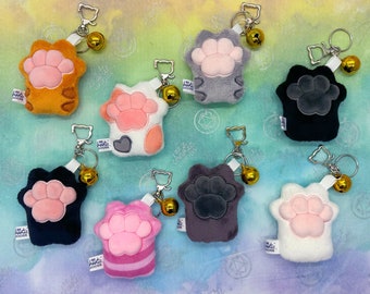 May include: Ten plush cat paw keychains with a bell charm. Each keychain features a different color and pattern, including orange, white, gray, black, pink, and purple. The keychains have the text "I'm a Magic  Doofus" printed on them.