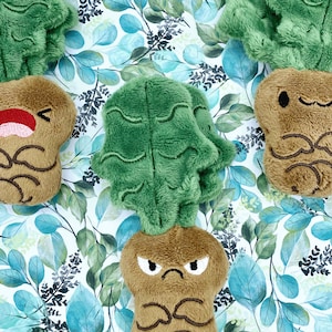 Puede incluir: Tres juguetes de peluche con forma de nabo con hojas verdes. Los nabos tienen diferentes expresiones faciales: uno sonríe, otro frunce el ceño y otro está sorprendido. Los juguetes están sobre un fondo blanco con un patrón de hojas verdes.