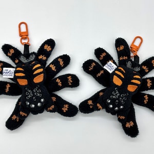 Peut inclure: Deux porte-clés en forme d'araignée en peluche noire avec des accents orange et le texte "The Rustic Goods" au dos.