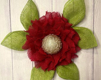 May include: A decorative flower with a red burlap center and green burlap leaves. The flower's center is a gold-colored sphere covered in glitter. The leaves are arranged around the flower in a starburst pattern.