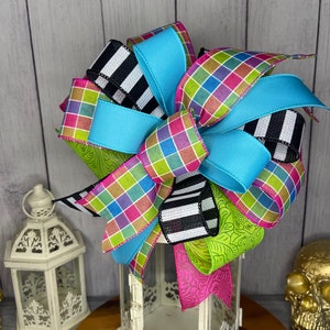 May include: A colorful bow made with black and white striped ribbon, turquoise ribbon, and a plaid ribbon with pink, green, and blue squares. The bow is tied around a white lantern.