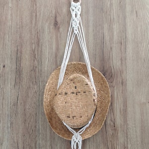 May include: A white macrame plant hanger with a wooden ring at the top. The hanger is hanging from a wooden surface and has a straw hat hanging from it.