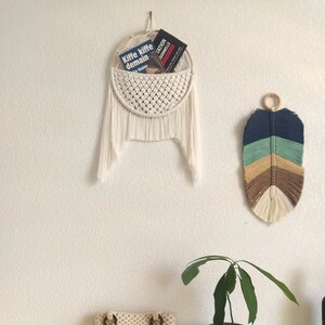 Peut inclure: Une suspension murale en macramé blanche avec une poche pour les livres. La suspension est décorée de glands blancs. Un petit sac en macramé blanc avec des anses en bois se trouve sur une table blanche. Un petit plant d'avocat en pot et un porte-encens en bois se trouvent également sur la table.