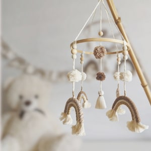 Könnte beinhalten: Ein Holz-Babymobil mit einem runden Rahmen und hängenden Dekorationen. Die Dekorationen umfassen weiße Pompons, braune Pompons und zwei gewebte Regenbögen mit weißen Fransen.