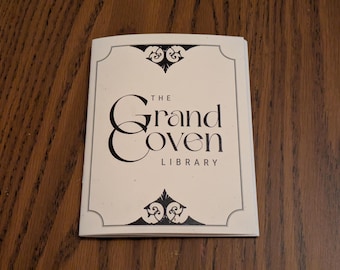 Op de afbeelding: Een crèmekleurig boekje met de tekst "THE Grand Coven LIBRARY" in zwarte, sierlijke letters. Het boekje heeft decoratieve zwarte krullen aan de boven- en onderkant. Het boekje ligt op een donker houten oppervlak.