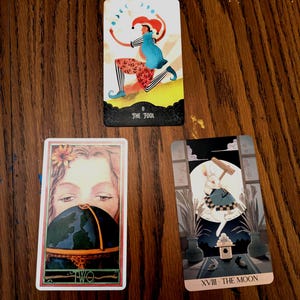 May include: Three tarot cards on a wooden surface. The top card features a jester with the text "THE FOOL". The second card shows a woman's face and a globe. The third card depicts a rabbit with the text "THE MOON".