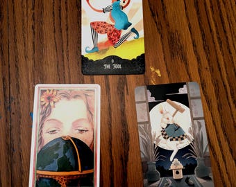 Op de afbeelding: Drie tarotkaarten op een houten oppervlak. De bovenste kaart toont een nar met de tekst "THE FOOL". De tweede kaart toont een vrouwengezicht en een wereldbol. De derde kaart toont een konijn met de tekst "THE MOON".