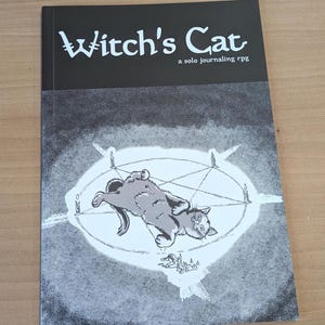 May include: A black and white illustration of a cat lying on its back inside a pentagram with five candles around it. The text "Witch's Cat" is at the top of the image, with the words "a solo journaling rpg" below it.