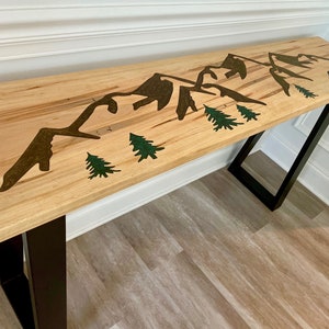 May include: A wooden console table with black metal legs features a mountain range and pine tree design. The table top is light brown with a natural wood grain.