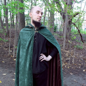 May include: A forest green cloak with a brown lining. The cloak has a hood with gold trim and a clasp closure. The person is wearing a black shirt and pants with brown boots. The cloak is made of a soft, velvet-like material.