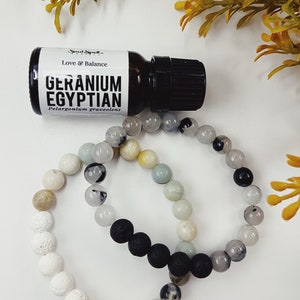 May include: A small bottle of essential oil labelled "Geranium Egyptian" with the text "Love & Balance" and "Pelargonium graveolens". The bottle is next to two bracelets made of white, black, and green beads.