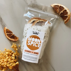 Puede incluir: Una bolsa de plástico transparente llena de sales de baño blancas, rodajas de cítricos secas y hierbas verdes. La bolsa tiene una etiqueta que dice "Soul Spell Apothecary Moon Spice Citrus + Mint All-Natural Vegan Bath Salt".