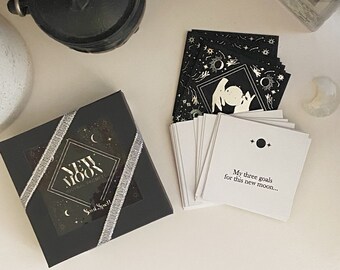 Op de afbeelding: Een zwarte doos met een zilveren lint en een transparant venster dat een zwart-witte illustratie van een wassende maan met sterren laat zien. De tekst "New Moon Journal Prompts" is in goud op de doos gedrukt. Een stapel witte kaarten met zwarte inkt en een zwart-witte illustratie van een wassende maan met sterren. De tekst "Mijn drie doelen voor deze nieuwe maan..." is op de bovenste kaart gedrukt.