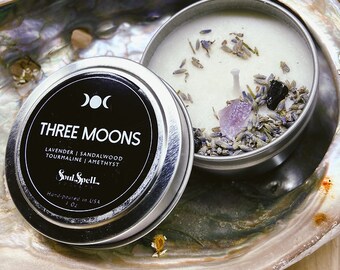 May include: A small, silver tin candle with a white wax top. The candle is decorated with dried lavender, amethyst crystals, and black tourmaline. The tin has a black label with the text "THREE MOONS" and "LAVENDER | SANDALWOOD TOURMALINE | AMETHYST" and the logo "Soul Spell".