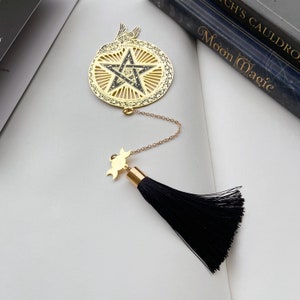 Puede incluir: Marcapáginas de oro con un pentagrama rodeado de una serpiente, un amuleto de luna creciente y un borla negra.