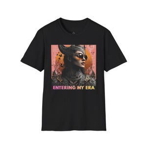 Pode incluir: Camiseta preta com um gráfico de uma mulher com óculos de sol e um capacete com chifres. A imagem tem um fundo rosa e laranja com o texto "ENTERING MY ERA" em gradiente.