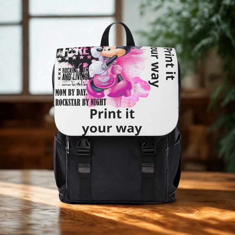 May include: A black backpack with a white flap featuring a graphic of a cartoon mouse playing a pink guitar. The text reads: Rocking and living, Mom by day, Rockstar by night, and Print it your way. The bag has two black buckle straps on the front.