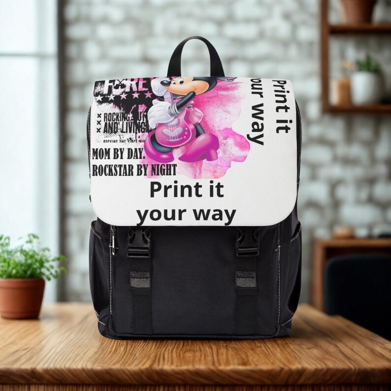 May include: A black backpack with a white flap featuring a graphic of a cartoon mouse playing a pink guitar. The text reads: FOREVER ROCKING AND LIVING, MOM BY DAY, ROCKSTAR BY NIGHT, and Print it your way. The bag has two black buckle straps.