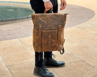 May include: A brown leather backpack with a distressed finish. The backpack has multiple pockets, adjustable straps, and metal buckles. The bag is held by a person wearing a white shirt and black trousers.