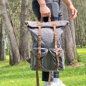 May include: A charcoal grey canvas roll-top backpack featuring two front pockets, brown leather straps with antique brass-toned metal buckles, and a top handle. The bag has a structured design with side pockets and reinforced leather detailing.