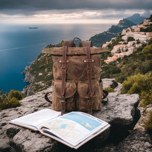 May include: A brown waxed canvas backpack with leather straps and accents sits on a rocky surface next to an open map. The backpack has two front pockets and a roll-top closure. The background features a coastal landscape with a town and the ocean.