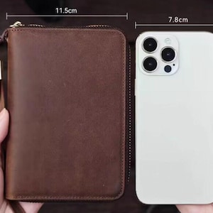May include: A brown leather wallet with a zipper closure and a gold pull tab. The wallet is 11.5 inches wide and 16 inches tall. A white smartphone with a camera lens is next to the wallet. The smartphone is 7.8 inches wide and 16 inches tall.
