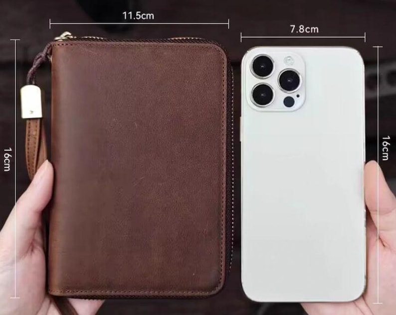 May include: A brown leather wallet with a zipper closure and a gold pull tab. The wallet is 11.5 inches wide and 16 inches tall. A white smartphone with a camera lens is next to the wallet. The smartphone is 7.8 inches wide and 16 inches tall.